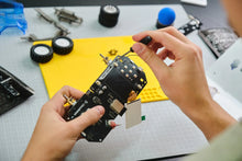 Load image into Gallery viewer, A close-up of a person's hands assembling a black electronic circuit board with gold connectors and a small antenna, working over a yellow grid mat on a gray workbench.