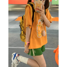 Load image into Gallery viewer, Person wearing an orange t-shirt with a graphic design, green shorts, and white sneakers on a street.