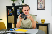 Load image into Gallery viewer, Person holding a camera lens in a room with a bookshelf and decorative items.
