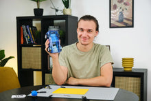 Load image into Gallery viewer, Person holding a blue electronic device in a room with a bookshelf and decorative items.
