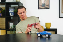 Load image into Gallery viewer, Man playing with a red gaming controller and blue toy truck in a room with shelves and decor.
