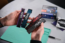 Load image into Gallery viewer, Three VSVN branded wristbands held in hands over a green work mat, with a product box and accessories on a gray table in the background.
