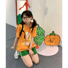 Load image into Gallery viewer, Child wearing an orange shirt with a cartoon design, sitting on the floor in front of a colorful wall decoration.
