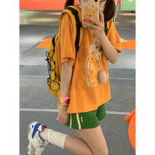 Load image into Gallery viewer, Person wearing an orange t-shirt with a graphic design, green shorts, and white sneakers on a street.
