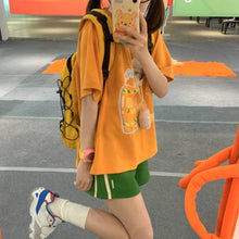 Load image into Gallery viewer, Person wearing an orange shirt with a cartoon character, green shorts, and white sneakers in an outdoor setting.
