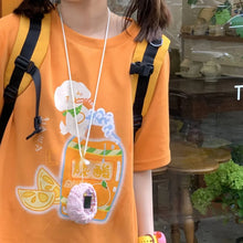Load image into Gallery viewer, A person wearing a bright orange graphic T-shirt with a cartoon drink design, a black-and-yellow backpack, and a pink fuzzy phone strap with a small screen, photographed in a real indoor setting by a window.
